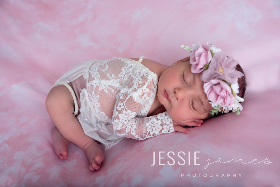 baby in taco newborn pose, baby sleeping with body laying over her legs with a floral headband on a pink backdrop