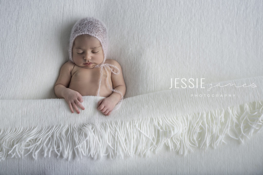 sleepy time newborn pose, baby in pink bonnet tucked in under a white blanket