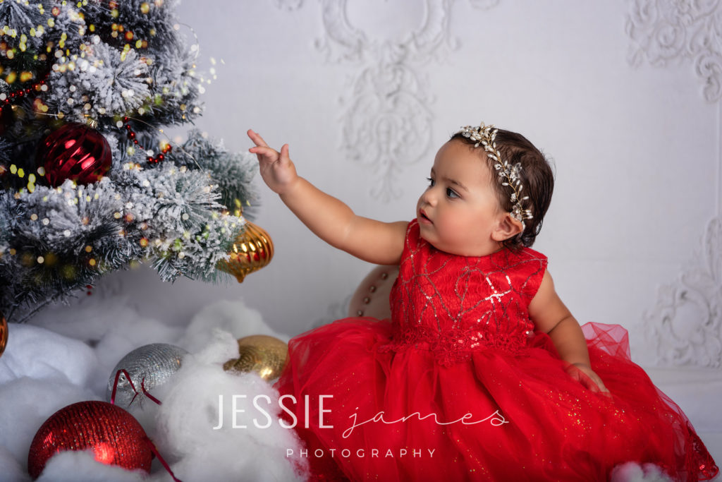 holiday mini session. toddler girl christmas photo, baby girl wearing a red dress touching a christmas tree