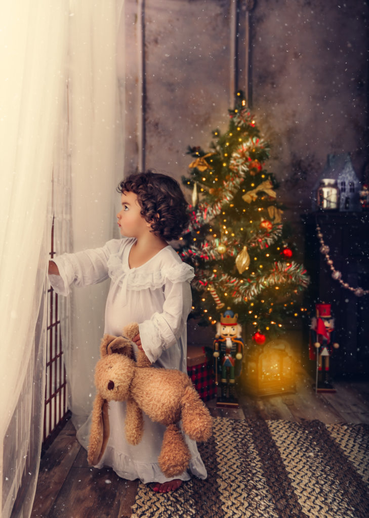 Chirstmas portrait of a little girl looking out the window with a white nightgown, holding a teddy bear.