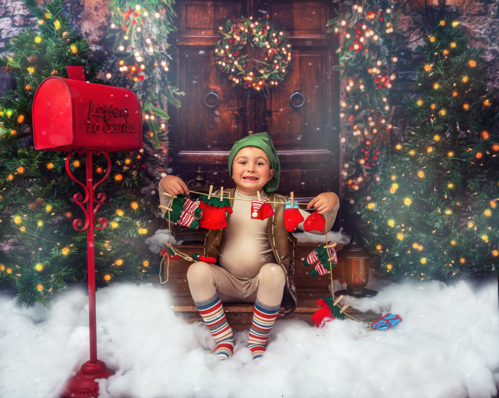 Little boy dressed as an elf sitting on stairs holding Christmas decorations.  Boy wearing green elf hat and striped socks.