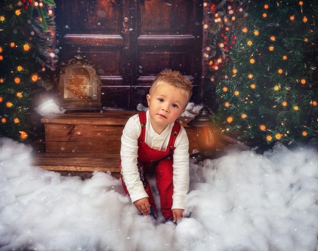 Little boy in red overalls sitting on a doorstep. Christmas portrait