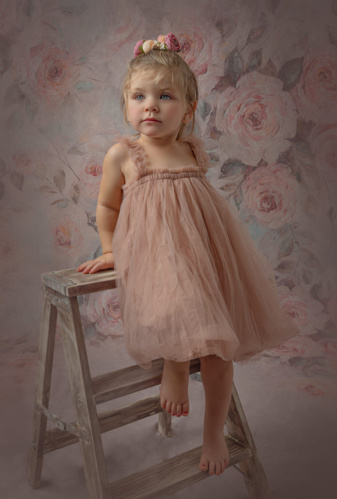 Little girl in a pink dress with blue eyes in front of a floral backdrop, leaning on a ladder. 