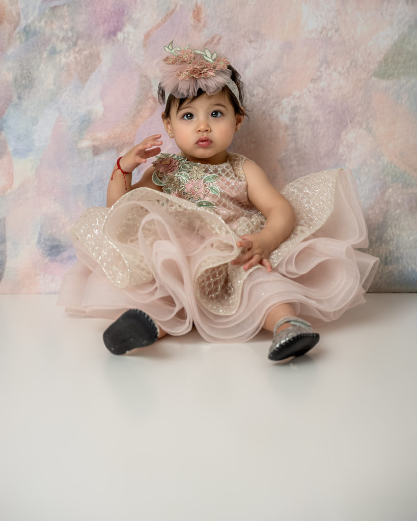 baby girl in a pink and gold dress sitting in front of a pastel colored backdrop. First birthday photo session
