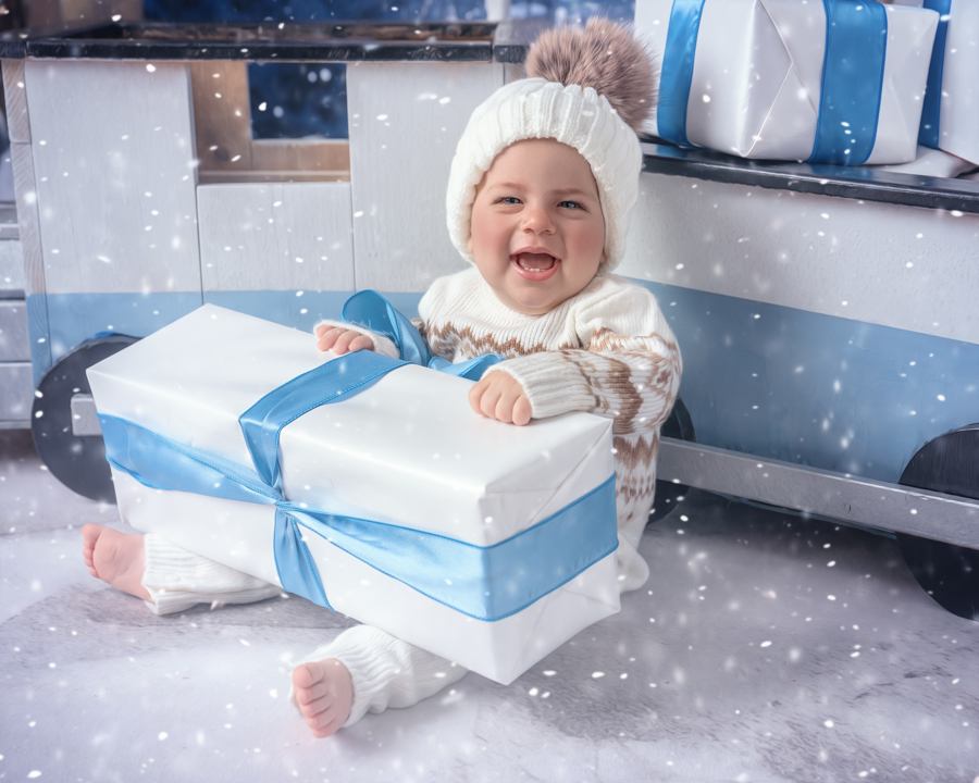 A baby boy sitting in front of a train with a present in the North Pole Express set, surrounded by snowy decorations and festive lights.