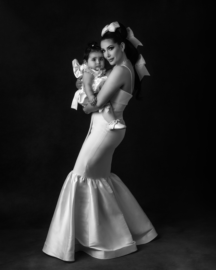 A mother in a form-fitting white satin gown holds her baby close, both adorned with elegant bows. The dramatic black background and sculpted lighting create a striking, fine-art portrait that feels timeless and editorial.