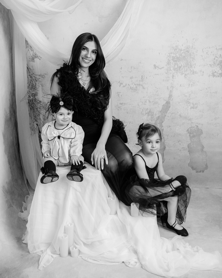 A mother sits gracefully with her two daughters, dressed in elegant black outfits, against a soft, neutral backdrop. This multi-generational portrait from a fine-art Mommy & Me session in New Jersey captures the beauty of family legacy.