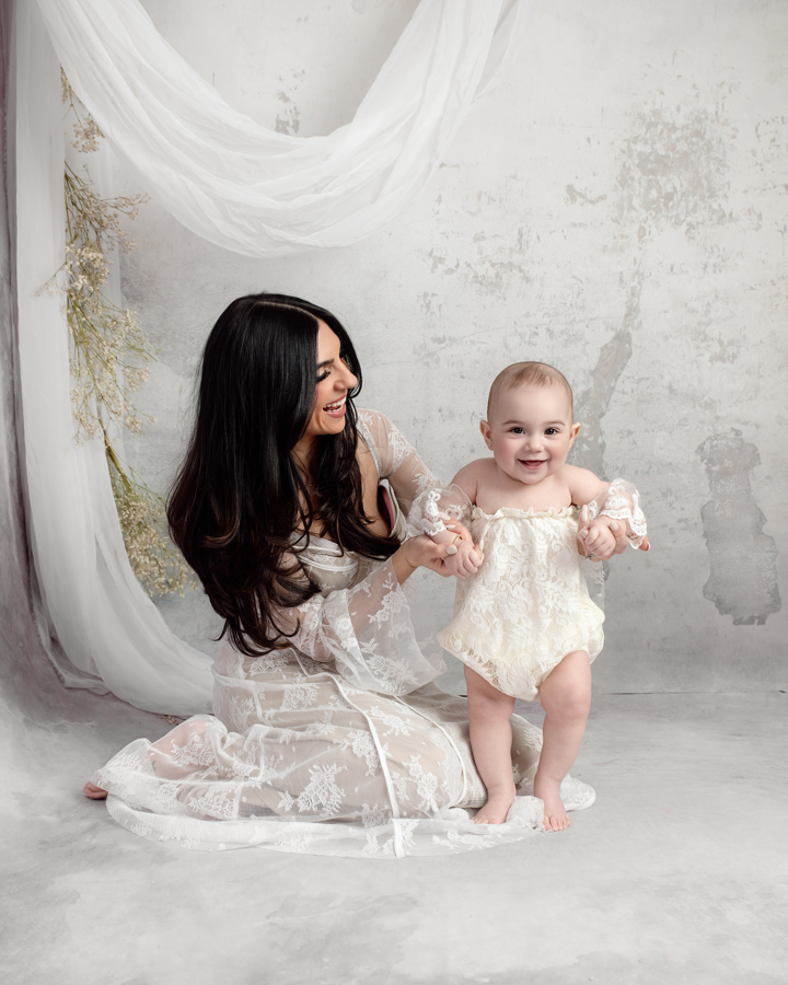 A joyful mother kneels on the floor, holding her baby’s hands as they share a playful moment. Captured in a Mommy & Me Mini Session in Staten Island, this fine-art portrait preserves the warmth and connection between mother and child.