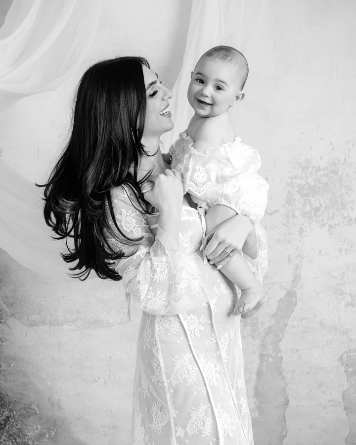 A mother in a lace gown holds her smiling baby against a soft, neutral-toned backdrop during a Mommy & Me Mini Session in Staten Island and New Jersey. The ethereal lighting and delicate textures create a fine-art motherhood portrait