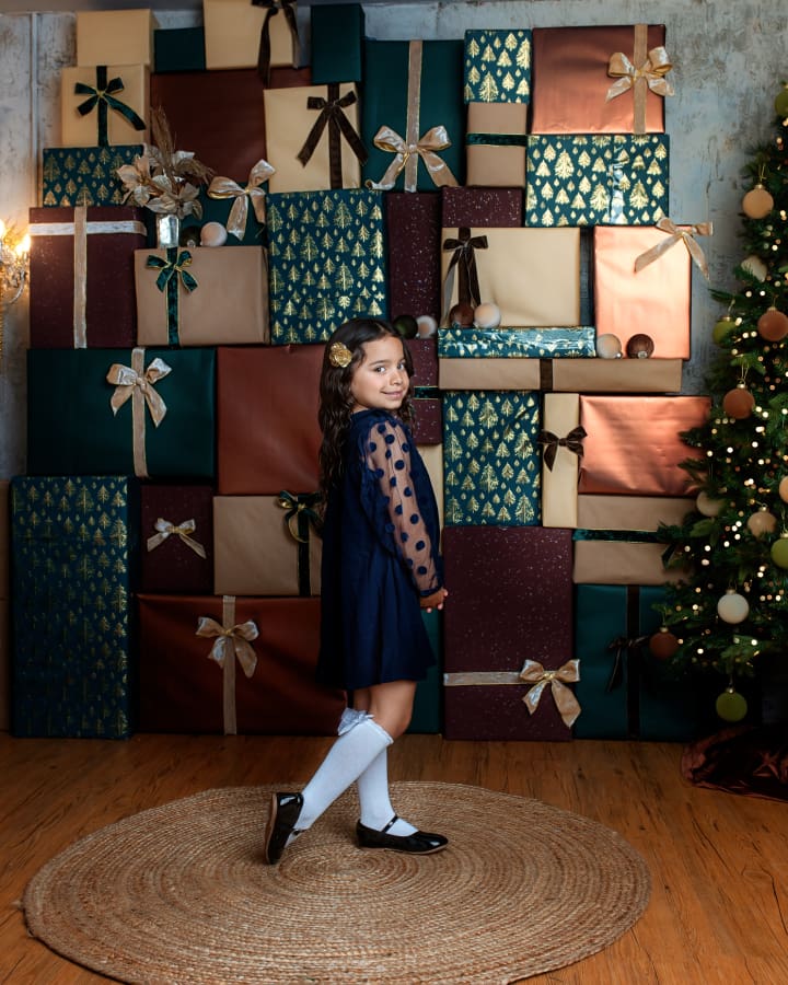 Smiling girl in navy holiday dress poses in front of stacked presents in The Holiday Gift Stack set