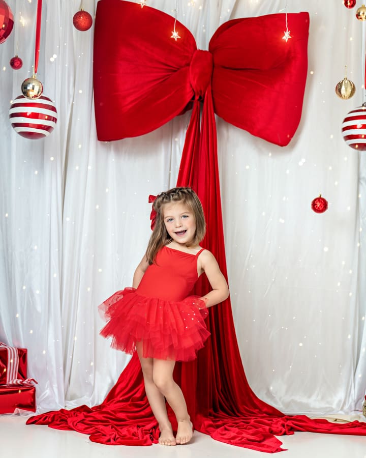 Little girl in a white holiday dress sitting with red presents under a giant red bow during Christmas Mini Sessions Staten Island.