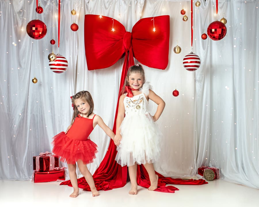 Two girls holding hands in front of oversized red bow and Christmas ornaments during Christmas Mini Sessions Staten Island.