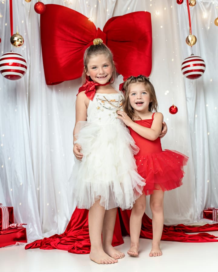 Little girl wearing a red tutu dress poses in front of a giant red velvet bow and Christmas ornaments during an Under the Mistlebow Christmas mini sessions in Staten Island studio.