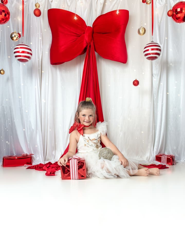 Smiling girl in red tutu dress poses with giant red bow and festive ornaments for Christmas Mini Sessions Staten Island.