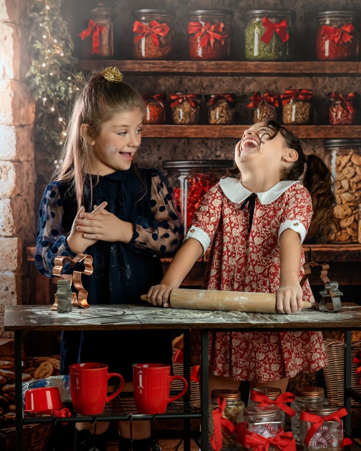 Two girls in vintage-style holiday dresses laugh together while baking during Christmas Mini Sessions Staten Island at the Christmas Cookie Cottage.