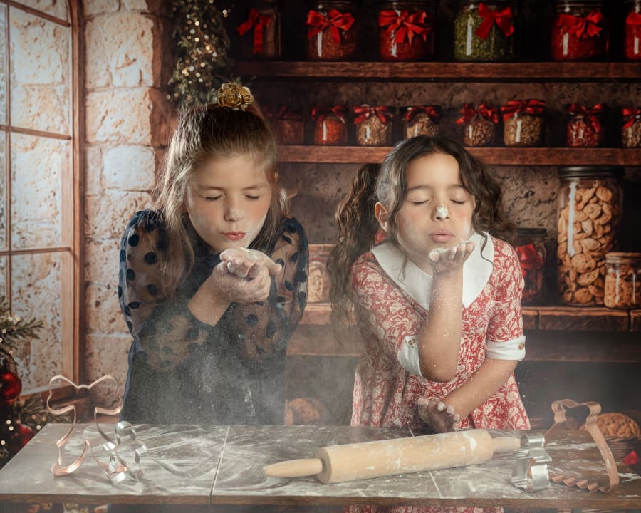 Two girls wearing vintage holiday dresses blow flour during Christmas Mini Sessions Staten Island in the Christmas Cookie Cottage set.