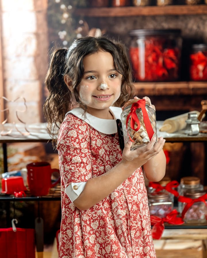 Smiling girl with flour on her face holds a jar tied with a red ribbon during Christmas Mini Sessions Staten Island at the Christmas Cookie Cottage.