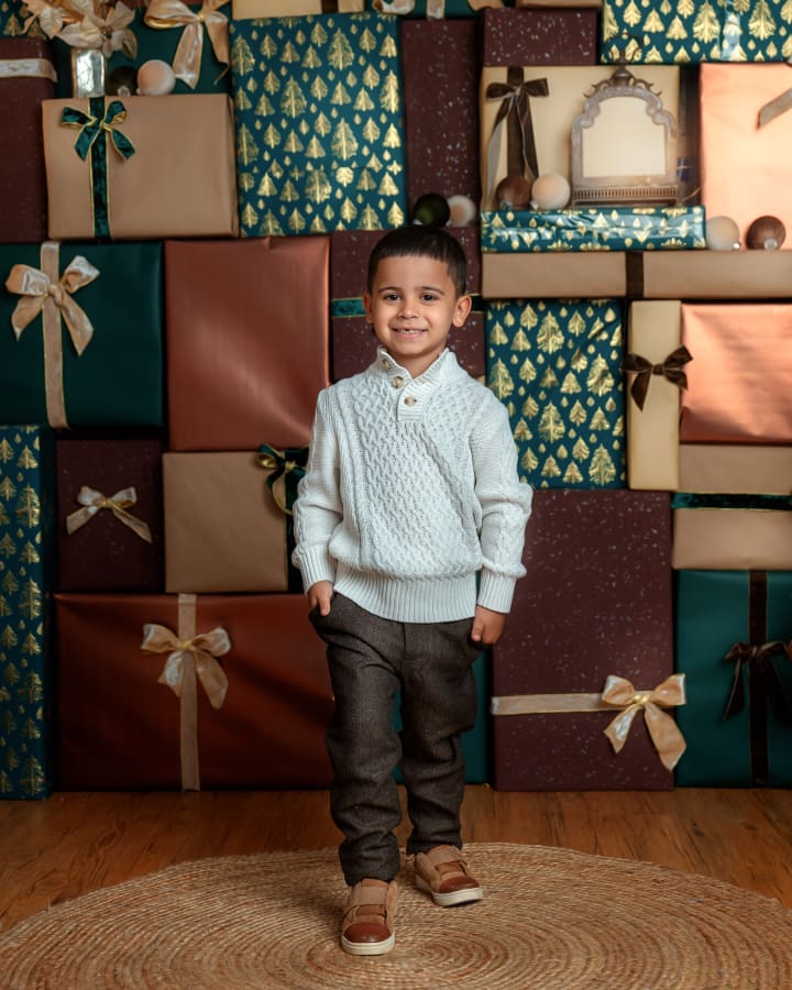 Young boy wearing a white sweater poses in front of colorful stacked gifts in The Holiday Gift Stack set during Christmas Mini Sessions Staten Island.